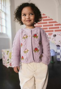 Jong kind met krullend haar dat een lila gebreide trui met bloemenborduursel en lichtbeige broek draagt, staande binnen bij een raam.