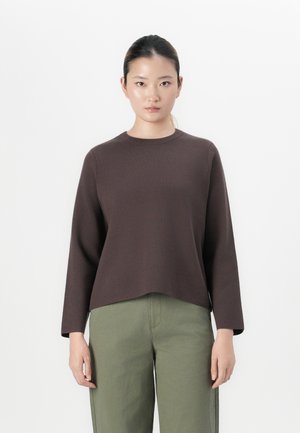 Woman stands facing forward wearing a dark brown long-sleeve top and olive green pants against a plain white background.