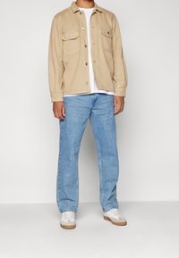 Beige oversized skjortjacka med två bröstfickor, bärs över en vit T-shirt, i kombination med ljusblå denimjeans och vita skor.