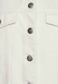 Casaco de ganga branca com botões de pressão metálicos, colarinho clássico e bolsos laterais, com uma textura subtil e pormenores de costura contrastantes.