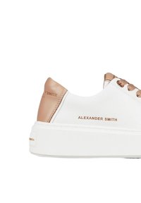 Sneaker bianco con tomaia in pelle liscia e dettagli marrone chiaro. Presenta lacci piatti beige, una suola testurizzata e il nome del brand impresso sul lato.