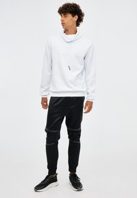 Sudadera blanca con un patrón de banda texturizada, cuello con cordón ajustable, combinada con pantalones jogger negros con detalles brillantes y puños elásticos. Zapatillas negras.