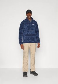 Temno modra pulover iz flisa z izbočeno vzorcem, ovratnik s polovičnim zadrgo in logotipom na prsih. Združeno s kremastimi cargo hlačami in črnimi pohodniškimi čevlji.