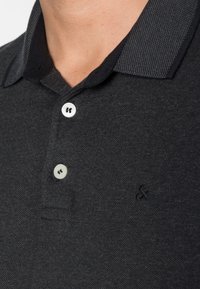 Musta polo-paita, jossa on teksturoitu kaulus, kaksi valkoista nappia ja pieni logo vasemmalla puolella. Kangas vaikuttaa pehmeältä ja teksturoidulta.