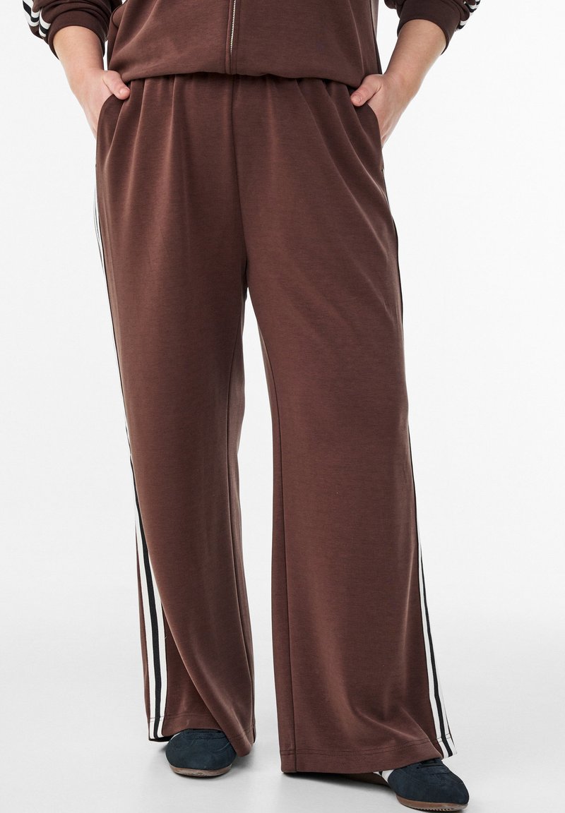 Person trägt braune Hose mit weitem Bein, weißen und schwarzen seitlichen Streifen, Hände in den Taschen und dunkle blaue Schuhe, steht vor weißem Hintergrund.