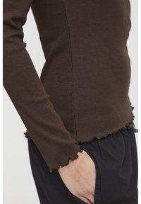 Braun geripptes Langarmshirt mit verkürztem Schnitt, das eine strukturierte Oberfläche und einen Rüschensaum aufweist. Kombiniert mit schwarzen, weit geschnittenen Hosen.