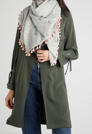 Femme portant un manteau vert foncé à manches longues avec des poignets noués, un jean bleu et une écharpe gris clair avec des pompons roses et de petites impressions noires et vertes.