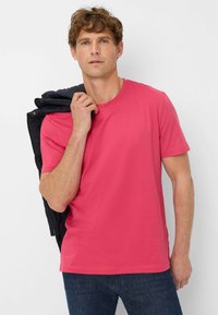 T-shirt a girocollo rosa realizzata in tessuto liscio, abbinata a jeans in denim scuri. Il modello tiene una giacca nera su una spalla.