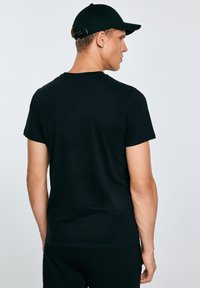 Férfi, aki egyszerű fekete pólót és fekete baseballsapkát visel, háttal áll, egyszínű világos háttér előtt.