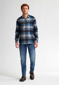 Camicia da uomo in flanella a quadri blu e grigi, con chiusura frontale a bottoni, maniche lunghe e due tasche sul petto. Abbinata a jeans blu e stivali marroni.