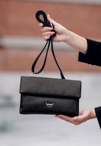 Černá kabelka z umělé kůže s překlopeným víkem, stříbrným logem a nastavitelným popruhem, držena v ruce proti rozmazanému pozadí.