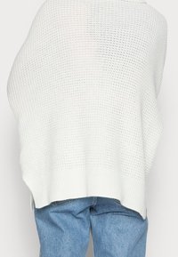 Suéter blanco de punto grueso y oversize con un diseño texturizado, que presenta un dobladillo acanalado, combinado con jeans de mezclilla azul claro.
