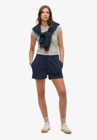 T-shirt gris avec graphique, short bleu marine avec poches latérales, et pull bleu marine noué autour des épaules. Baskets beiges et chaussettes claires.