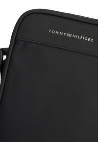 Λεπτομέρεια μαύρης τσάντας χιαστί Tommy Hilfiger που δείχνει το όνομα της μάρκας σε ασημένιο γράψιμο πάνω σε υφασμάτινη υφή με ομαλή επένδυση.