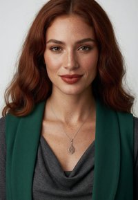 Sølvhalskæde med vedhæng formet som et blad, båret over en draperet grå top og fremhævet af et mørkegrønt tørklæde. Glatte teksturer og delikate detaljer.