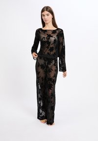 Schwarzes, transparentes Outfit mit langen Ärmeln und Hose, das mit gestickten floralem Mustern und einer taillierten Taille ausgestattet ist, wodurch eine fließende Silhouette entsteht.