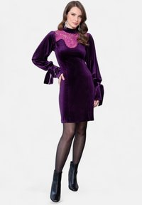 Abito in velluto viola con maniche lunghe, collo alto decorato con pizzo rosa e dettagli a fiocco sulle maniche. Abbinato a stivaletti neri alla caviglia.