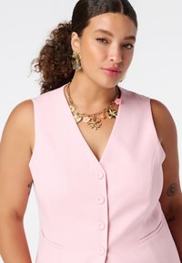 Roze mouwloze blazer met een diepe V-hals en vijf knopen aan de voorkant, gecombineerd met een gouden florale statement ketting en grote florale oorbellen.