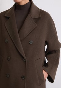 Brauner Wollmantel mit doppelter Knopfleiste, großen geschlitzten Revers, vier schwarzen Knöpfen und zwei seitlichen Taschen.