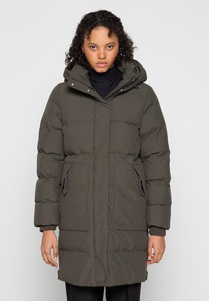 Jonge vrouw die een lange, olijfgroene gewatteerde winterjas met kap en ritssluiting aan de voorkant draagt tegen een effen lichte achtergrond.