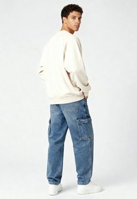 Sweat-shirt beige à coupe décontractée avec des poignets côtelés, associé à un jean cargo bleu clair avec poches plaquées. Des baskets blanches complètent le look.