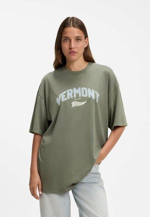 Vrouw met lang bruin haar die een oversized olijfgroen t-shirt draagt met de tekst "VERMONT" en lichtblauwe spijkerbroek, staand tegen een witte achtergrond.