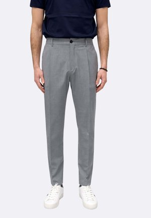 Uomo che indossa pantaloni aderenti grigio chiaro, sneakers bianche, braccialetto nero con perline e camicia a maniche corte blu scuro, in piedi su sfondo bianco.