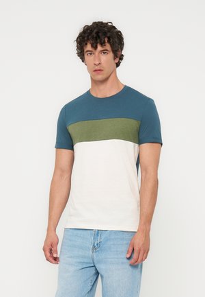 Mężczyzna z kręconymi włosami, ubrany w krótkim rękawem T-shirt w blokach kolorów niebieskiego, zielonego i białego oraz jasnoniebieskie dżinsy, stojący na białym tle.