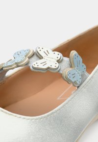 Γκρι ανοιχτό παπούτσι με ομαλή υφή, διακοσμημένο με λουράκι που φέρει μπλε και λευκές ξύλινες πεταλούδες. Μαλακό μπεζ εσωτερικό.