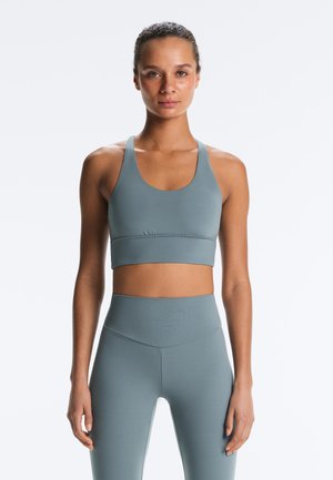 Jonge vrouw draagt een bijpassende blauwgrijze sportbeha en hoge taille legging, staande met gezicht naar voren tegen een witte achtergrond.