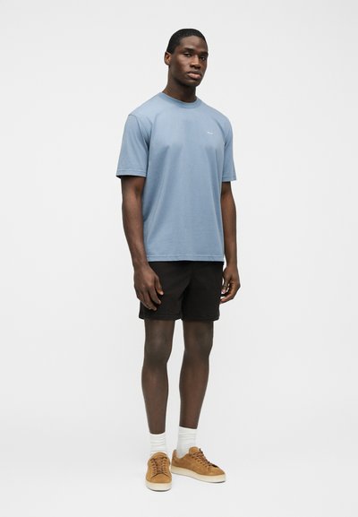 Homme portant un t-shirt bleu à col rond, un short noir, des chaussettes blanches et des baskets beige, debout devant un fond blanc uni.