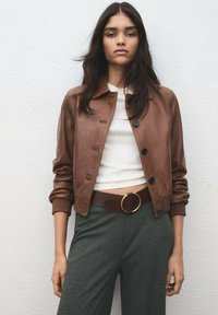 Brun skinnbomberjacka med krage, ribbade ärmslut och knäppning framtill; kombinerad med grå byxor och ett brunt bälte med en rund spänne.