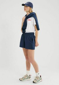 Marineblaue Shorts mit elastischem Bund, kombiniert mit einem weißen T-Shirt mit rotem Logo und einem marineblauen Pullover über den Schultern. Sportschuhe.
