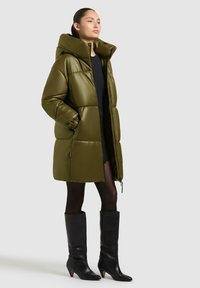 Olive green puchowy płaszcz z wysokim kołnierzem, z zamkiem z przodu i elastycznymi mankietami. Noszony na czarnej sukience i zestawiony z botkami za kolano.