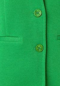 Grüne Blazerjacke aus strukturiertem Stoff, mit zwei großen grünen Knöpfen und einer kleinen Tasche auf der linken Seite. Einfaches und strukturiertes Design.