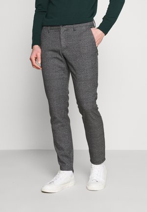 Mann trägt graue karierte Slim-Fit-Hose, weiße Sneakers und ein dunkelgrünes Langarmshirt, steht mit einer Hand in der Tasche.