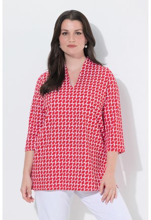 Donna con capelli lunghi e scuri che indossa orecchini di perle, una camicetta rossa e bianca a motivi con maniche a tre quarti e pantaloni bianchi.