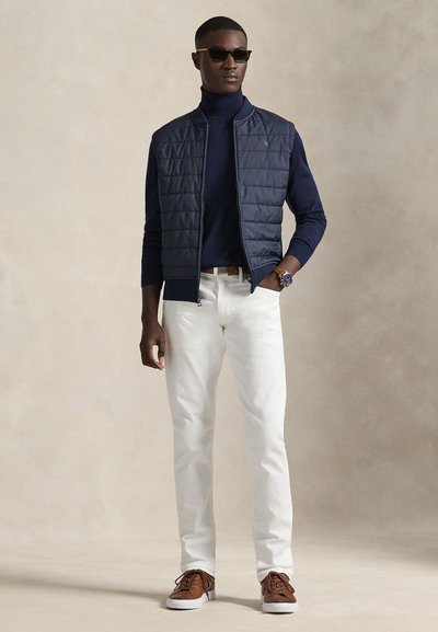 Veste matelassée bleu marine portée sur un col roulé foncé, associée à un pantalon blanc et des baskets marron. Accessoires : lunettes de soleil et montre.