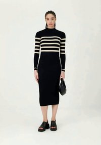 Ženska v črni midi obleki z bež vodoravnimi črtami na zgornjem delu in rokavih, ki drži črno torbico in nosi črne sandale na odprto konico.