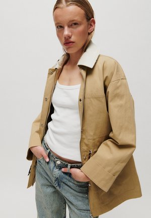 Frau trägt eine hellbraune Jacke mit weißem Lammfellkragen, weißes Tanktop und blaue Jeans, steht mit Händen in den Taschen vor einem einfarbigen Hintergrund.