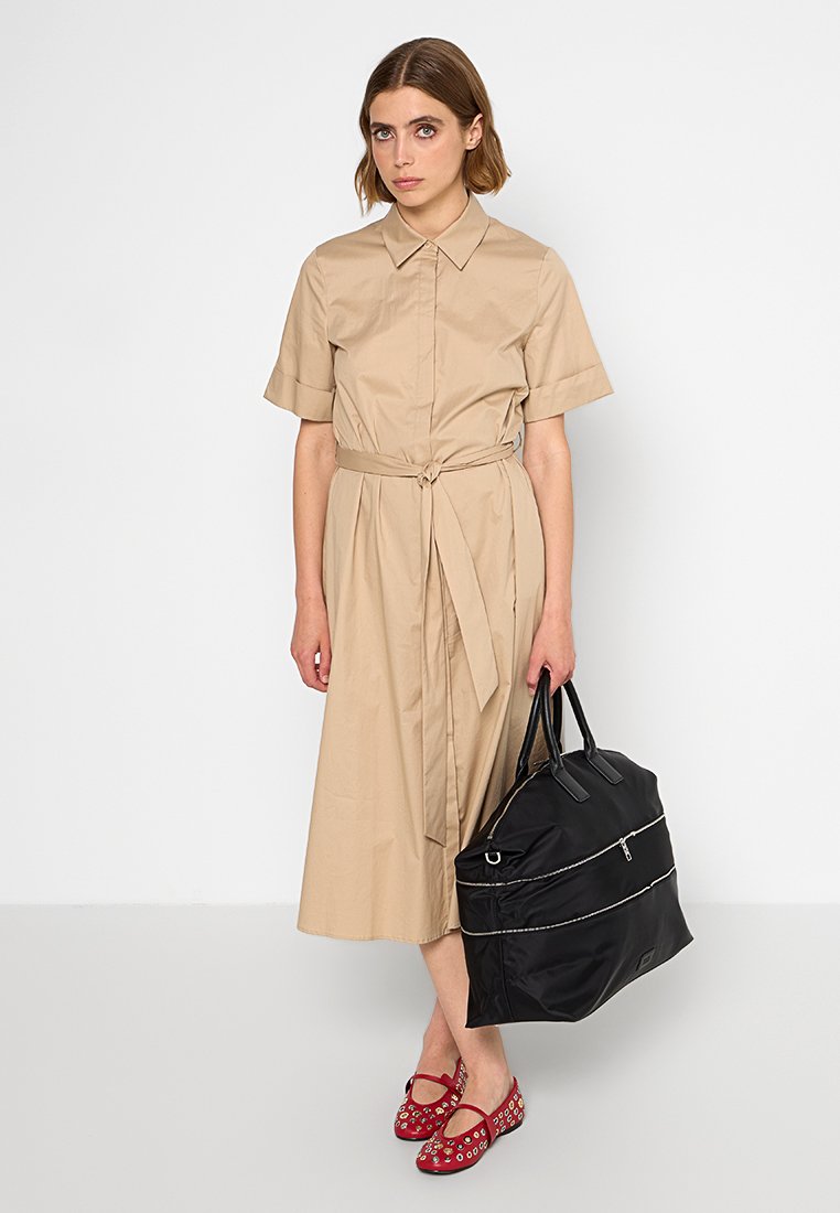 Vestido de camisa beige hasta la rodilla con mangas cortas, cintura atada, combinado con un bolso negro con detalles en plateado y zapatos rojos con tachuelas decorativas.