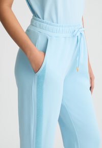 Pantaloni della tuta azzurro chiaro con vita elastica, tasche laterali e un dettaglio a righe testurizzate lungo le gambe esterne. Tessuto morbido.