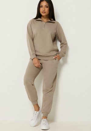 Mujer con sudadera beige con cremallera y pantalones deportivos a juego con zapatillas blancas, de pie junto a una pared blanca.