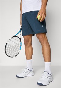 Pantalones cortos de tenis azul marino con bolsillos, calcetines de algodón blancos y zapatillas blancas con detalles en azul marino. Sosteniendo una pelota de tenis verde y una raqueta.