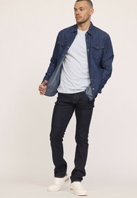 Chemise en denim bleu foncé avec poches poitrine sur un t-shirt bleu clair, associée à un jean foncé et des baskets blanches. Coupe décontractée, design à boutons.