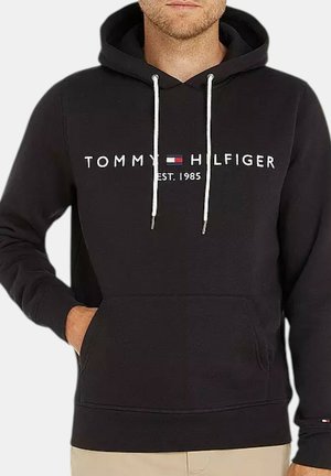 Mężczyzna noszący czarną bluzę z kapturem Tommy Hilfiger z białymi ściągaczami i logo z napisem „TOMMY HILFIGER EST. 1985” na piersi, ręka w kieszeni.