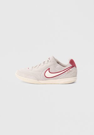 Zapatilla de ante gris claro con detalles en rojo, que presenta un prominente logo swoosh blanco y suela de goma texturizada. Diseño con cordones y cuello acolchado.