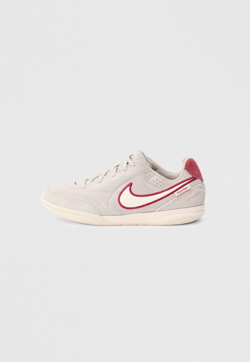 Lichtgrijze suède sneaker met rode accenten, voorzien van een opvallend wit swoosh-logo en een gestructureerde rubberen zool. Vetersluiting met gevoerde halslijn.