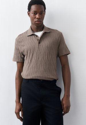 Junger Mann, der ein strukturiertes, beiges kurzärmeliges Poloshirt und schwarze Hose trägt, vor einem schlichten weißen Hintergrund stehend.