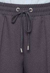 Pantalons de jogging gris foncé avec une taille à cordon de serrage, dotés d'œillets et de embouts en métal. Le tissu a une apparence douce et texturée.